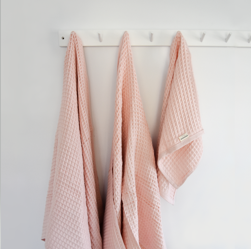 Two taffy pink children's organic cotton waffle towels and an organic cotton waffle hand towel hang on a white hanging rack on a wall.