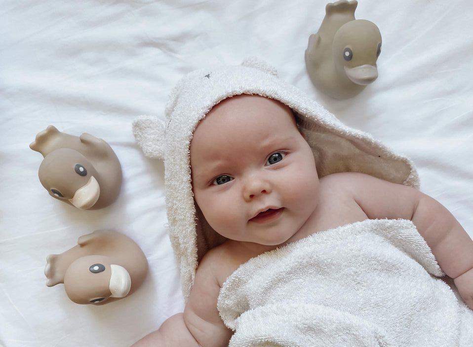 Baby wearing a hooded towel surrounded by 3 natural rubber duck bath toys