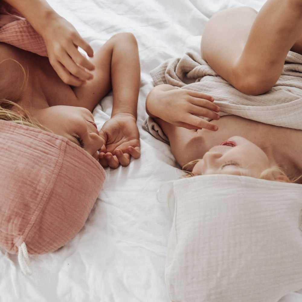 two girls lying on a bed wrapped in organic hooded cotton children's towel, one wears a neutral tan latte colour the other a dusk pink