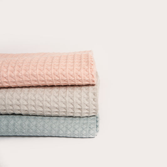 three organic waffle towels neatly folded in a stack. they are coloured pink light grey and blue green