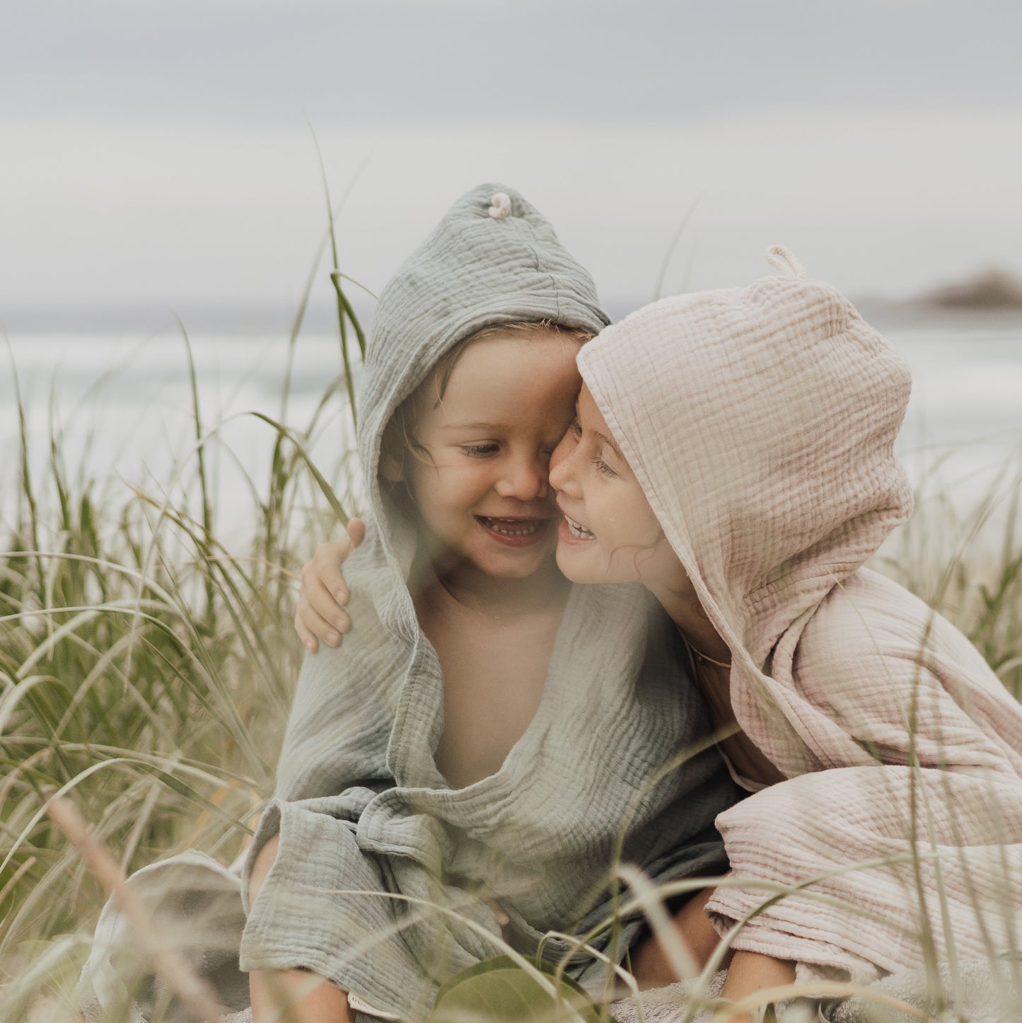 two children sitting and smiling while organic hooded cotton children's towel, one is wearing storm green, the other in neutral latte